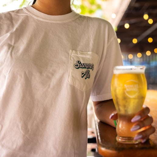 Front and back view of a white Samuel Adams t-shirt with “Summer Ale” text on the front pocket and a large, colorful summer-themed graphic on the back featuring a sun, palm tree, ocean waves, and the Samuel Adams logo.