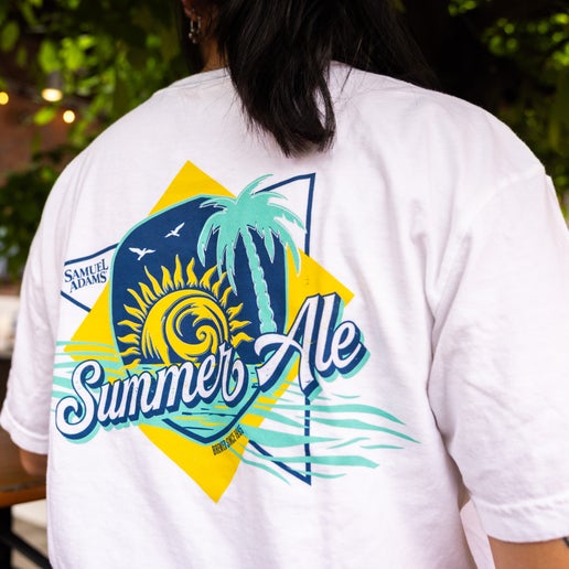 Front and back view of a white Samuel Adams t-shirt with “Summer Ale” text on the front pocket and a large, colorful summer-themed graphic on the back featuring a sun, palm tree, ocean waves, and the Samuel Adams logo.