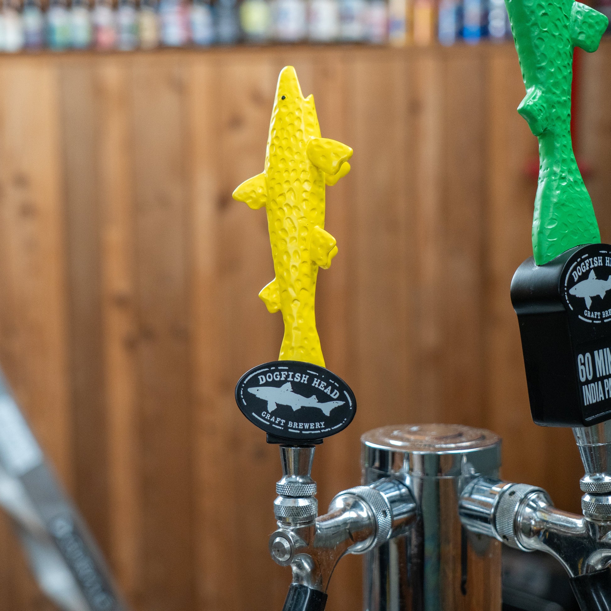 A neon yellow, shark-shaped short handle from Merchtank, the "Yellow Shark Tap Handle Shorty," adorns the beer tap, with a wooden wall and shelves of Dogfish beers blurred in the background.