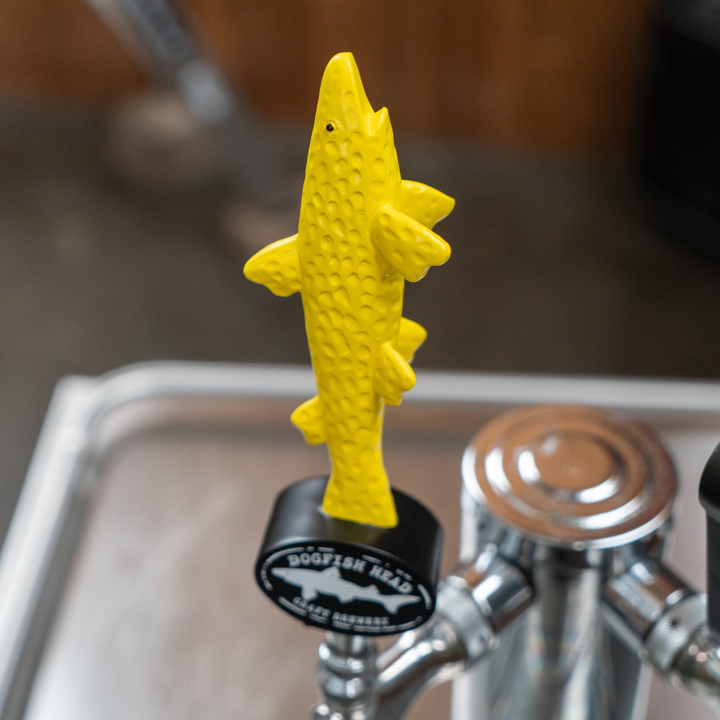 A vibrant neon yellow, shark-shaped short handle labeled Merchtank's "Yellow Shark Tap Handle Shorty" is attached to a beer tap, standing out boldly against the blurred backdrop of a bar or brewery setting—an ideal eye-catcher for serving Merchtank beers.