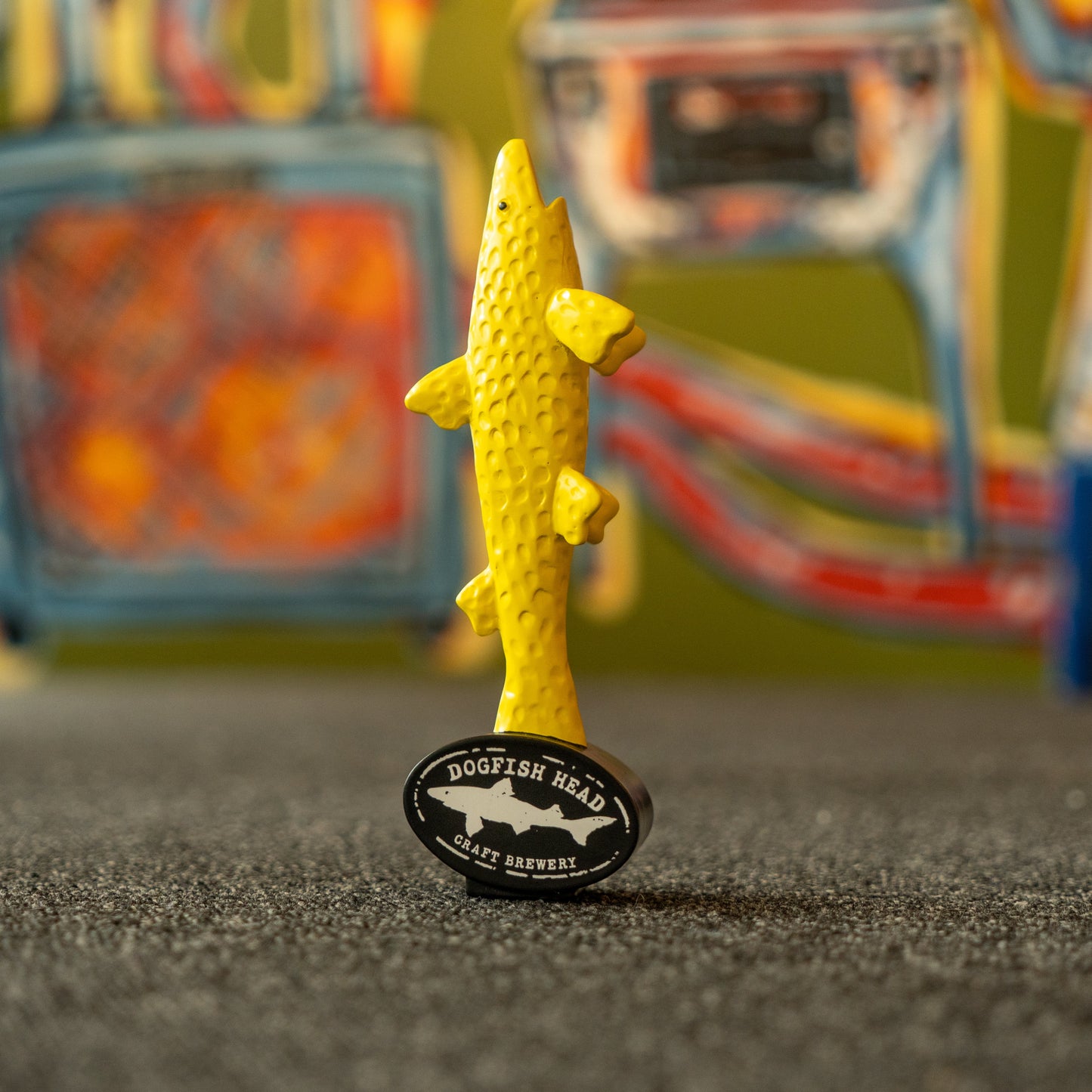 A vibrant neon yellow, fish-shaped short tap handle—the Yellow Shark Tap Handle Shorty by Merchtank—stands atop a textured surface, with colorful abstract art blurred in the background; a striking accent ideal for pouring your favorite beers.