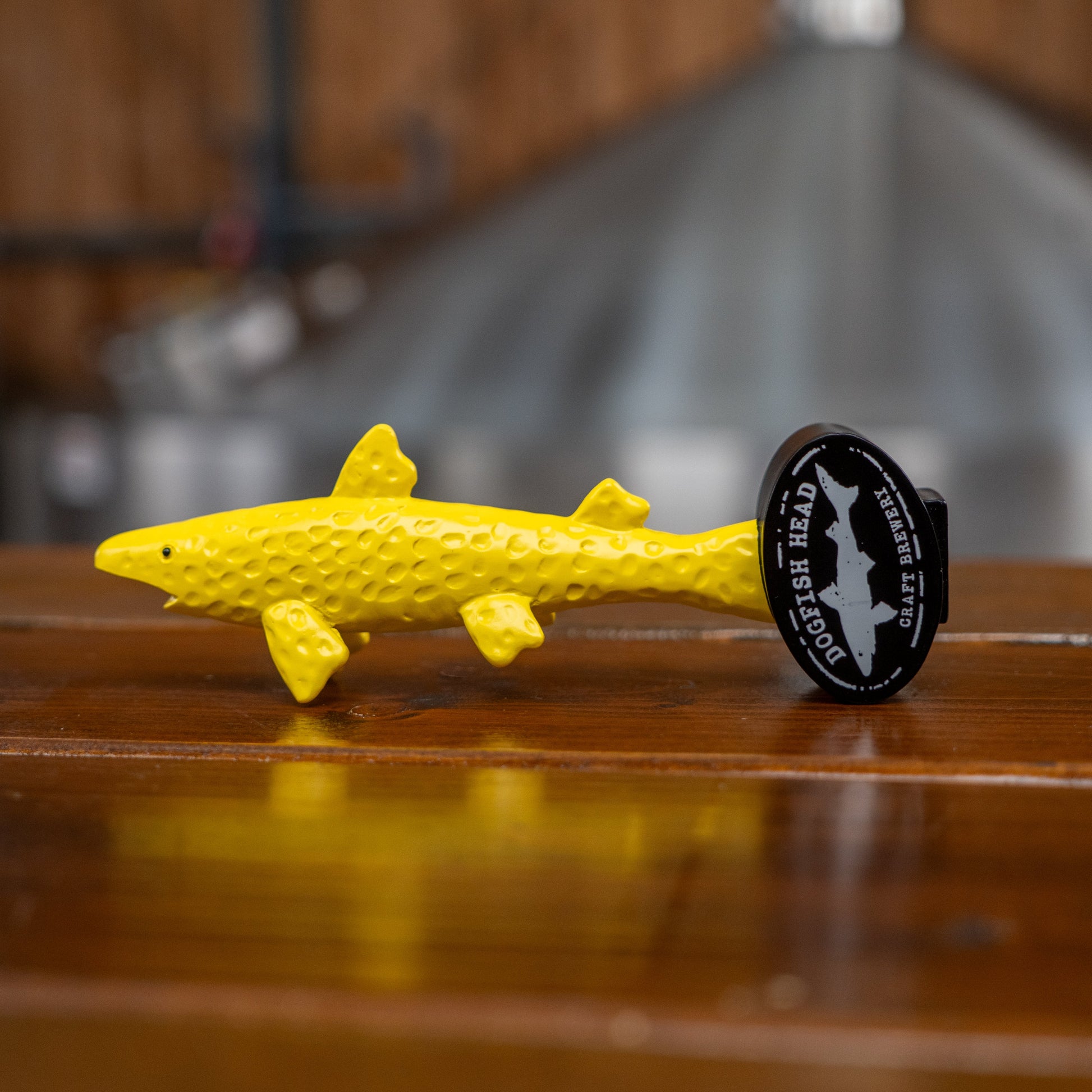 A neon yellow Yellow Shark tap Handle Shorty by Merchtank, shaped like a shark with a short handle, rests on a wooden surface. Near the tail, a circular Dogfish Head Craft Brewery logo is attached—perfect for opening your favorite Dogfish beers. The background is blurred.