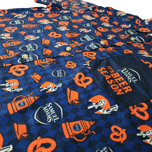 Close up of Blue button up short sleeve shirt designed with many small Samuel Adam shield logos, orange pretzels, and orange Octoberfest steins. Has a pocket on the left chest with a navy Samuel Adams shield logo and "It's BEER SEASON" in orange 