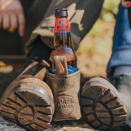 Samuel Adams x BLUNDSTONE Leather Can and Bottle Cooler on a Boston Lager bottle being held between a persons boots.