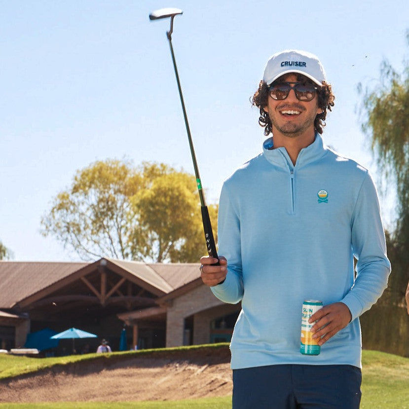 Man in light blue pullover and white cap holding a golf club and can on a golf course